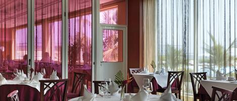Un restaurant avec des tables et des chaises rondes, de grandes fenêtres avec des rideaux et une vue sur des palmiers.