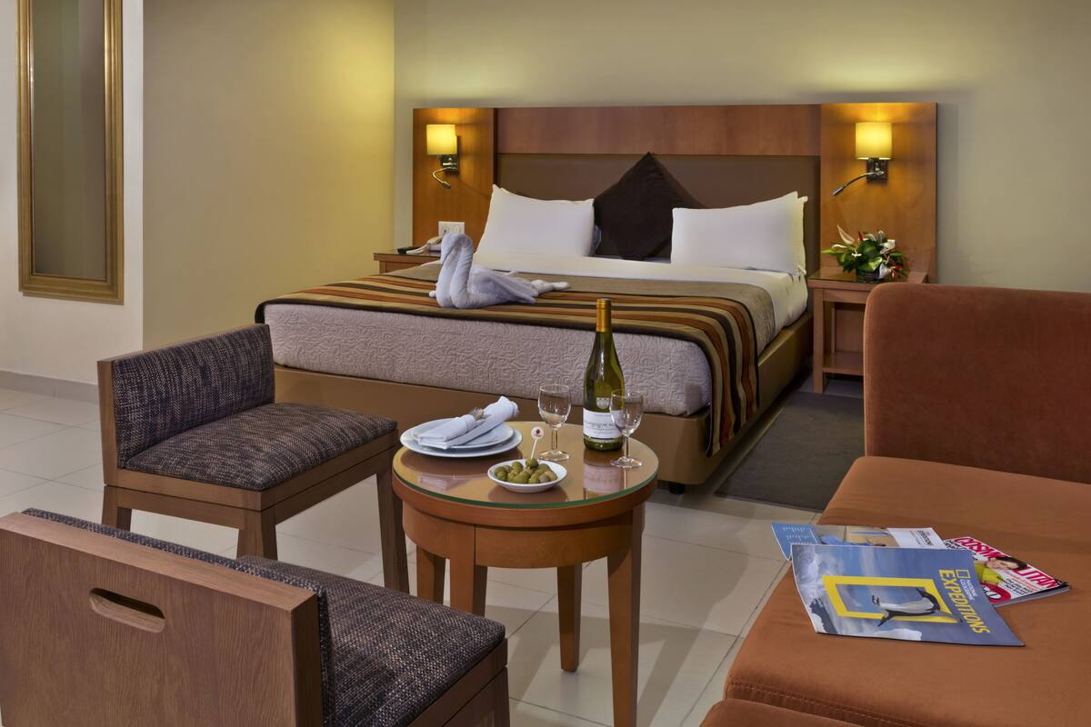 A hotel room with a bed, a dining table with a bottle of wine, a chair, a sofa, and a magazine.