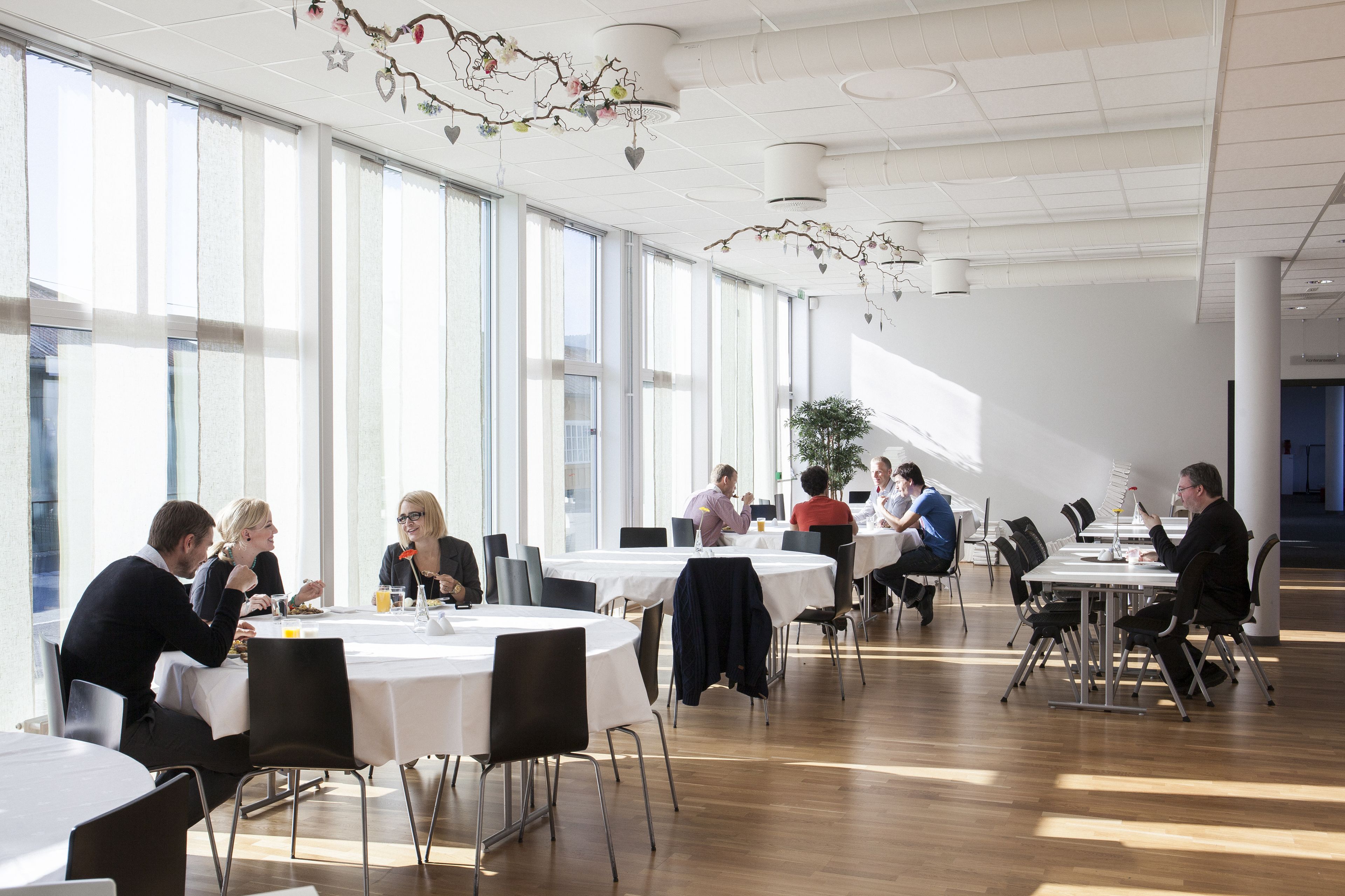 A spacious dining area with round tables and chairs, large windows, and a modern ceiling design.