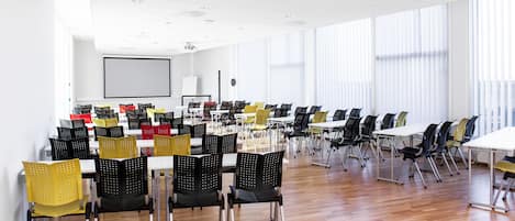 Un salón de conferencias con varias mesas y sillas, una pantalla de proyector y ventanas grandes con persianas.