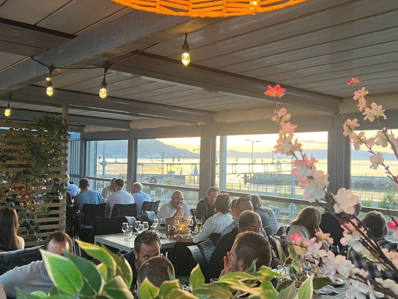 A restaurant with patrons seated at tables, large windows offering a view of a waterfront, and hanging light fixtures.