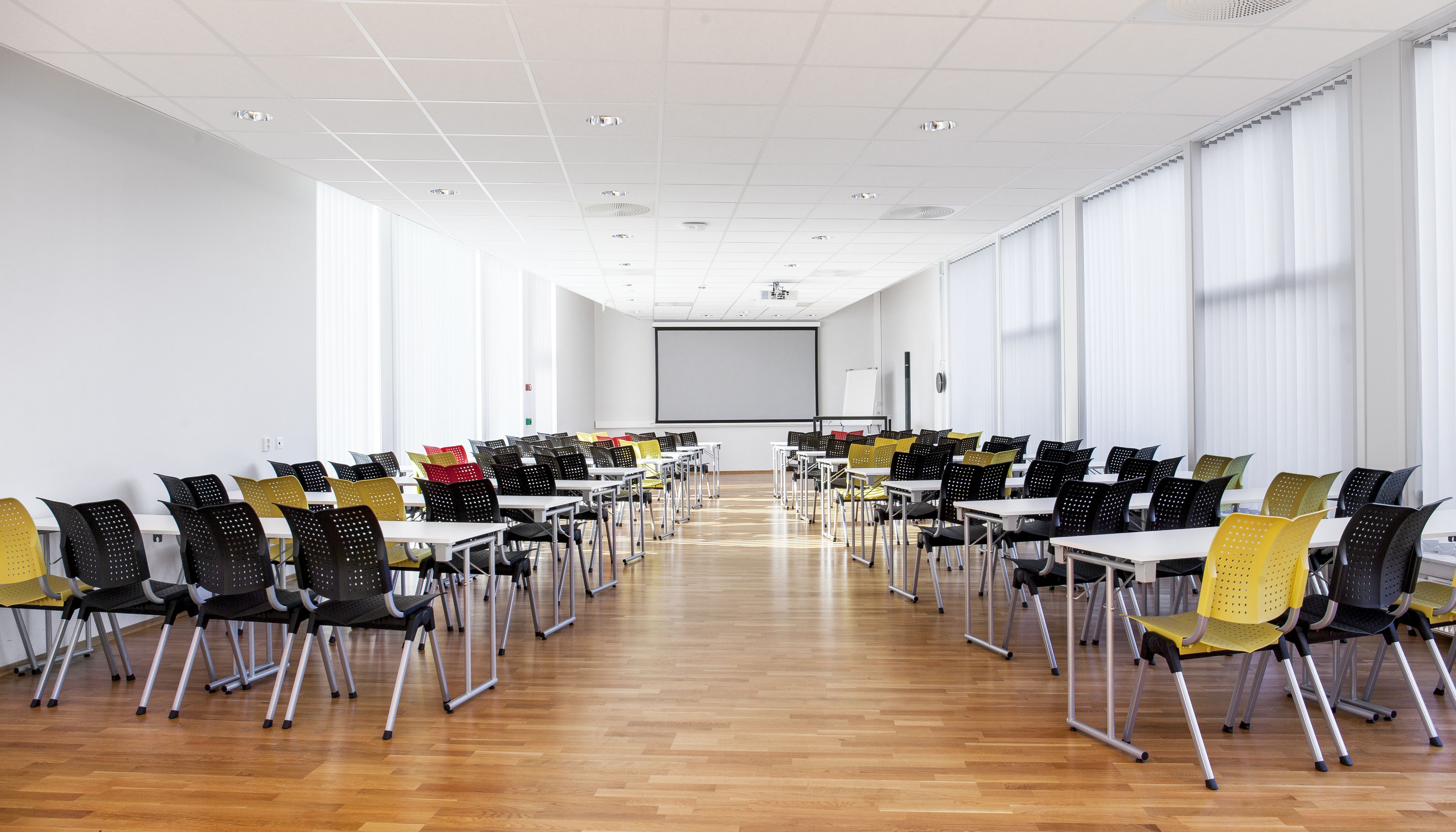 Un salón de conferencias con filas de mesas y sillas, ventanas amplias y una pantalla para proyector.
