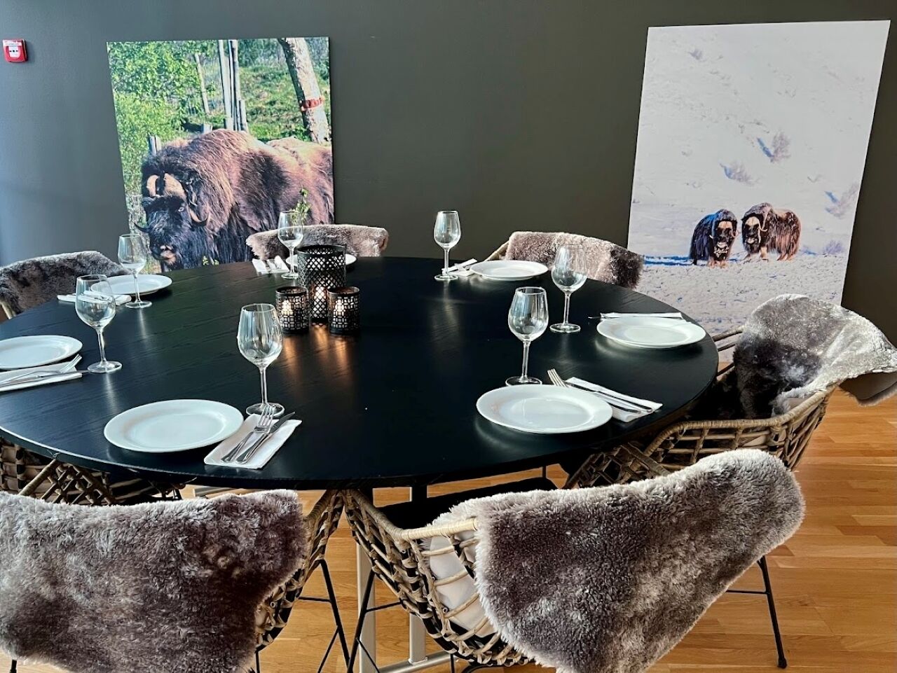 A round dining table set for six with plates, cutlery, and wine glasses. There are two framed pictures on the walls behind the table.