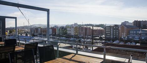 Una terraza de madera con mesas y sillas que da a un puerto deportivo con barcos y edificios al fondo.