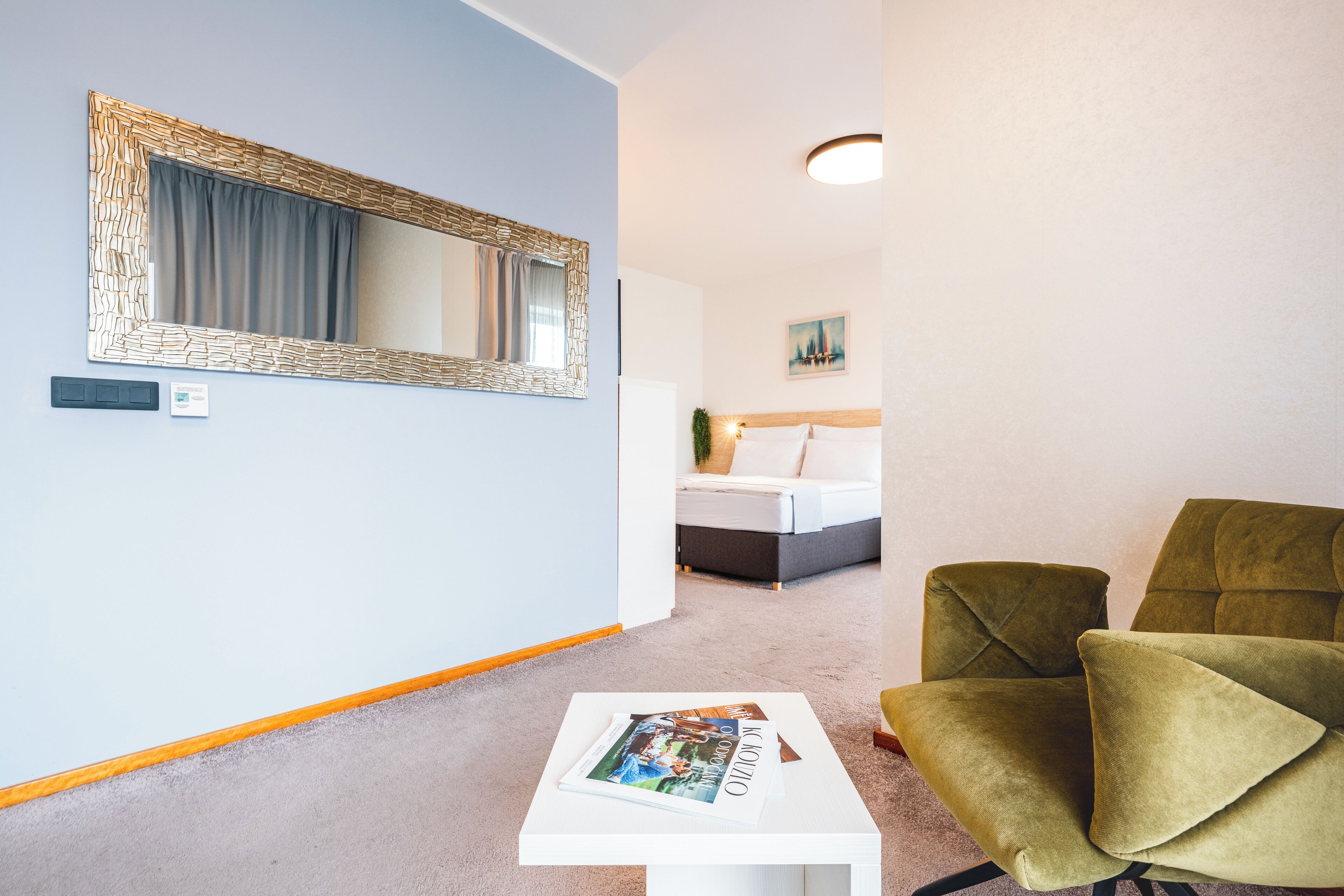 A room with a bed, a green armchair, a white table with a magazine, a large mirror, and a painting on the wall.