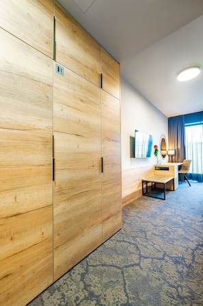 A hotel room with a wooden wardrobe, a desk with a chair, a small table, a TV on the wall, and a window with curtains. - Maximus Resort (Brno)
