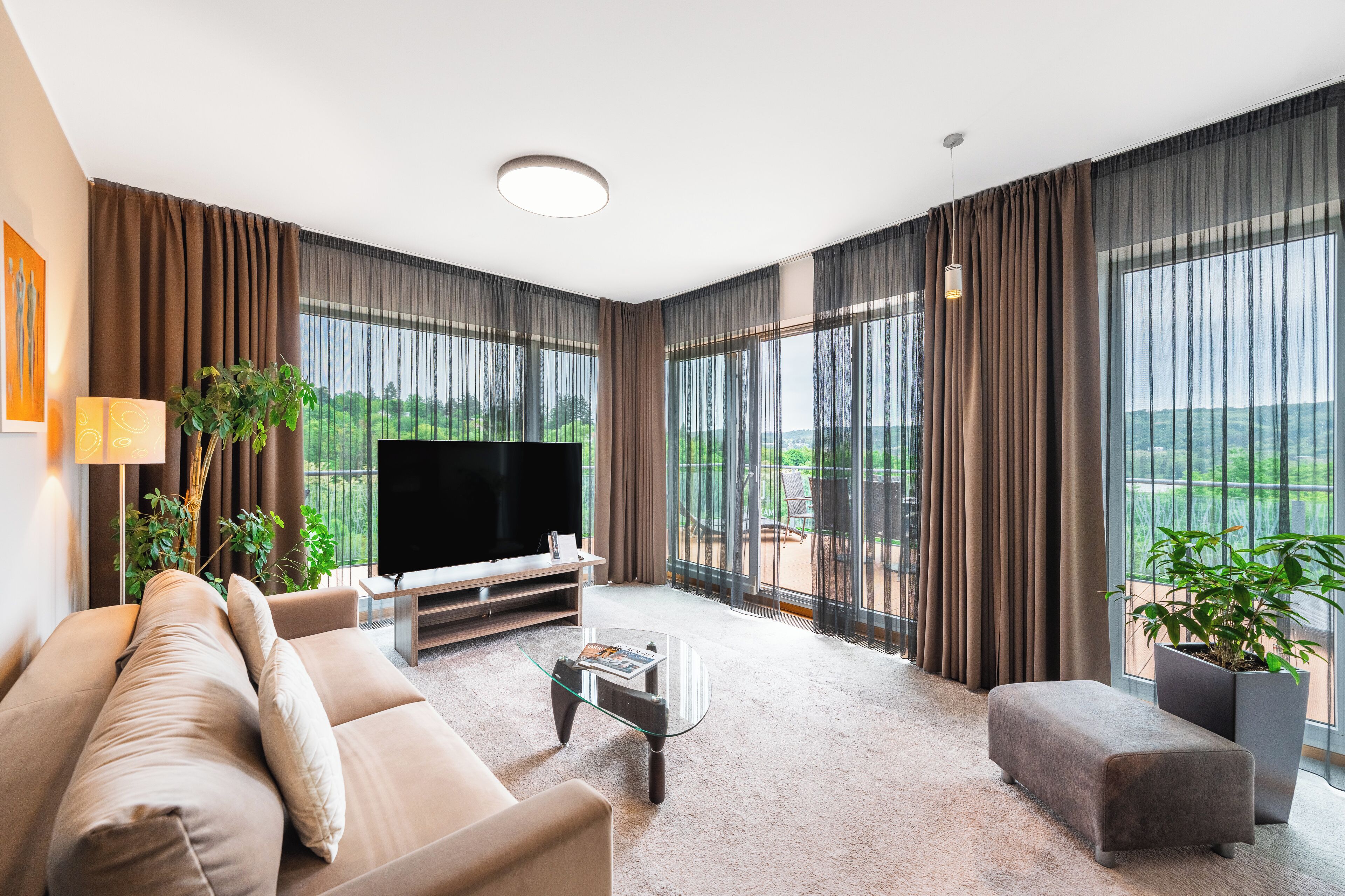 Una sala de estar moderna con sofá, mesa de centro y televisor, ventanas amplias con cortinas y vistas al balcón.