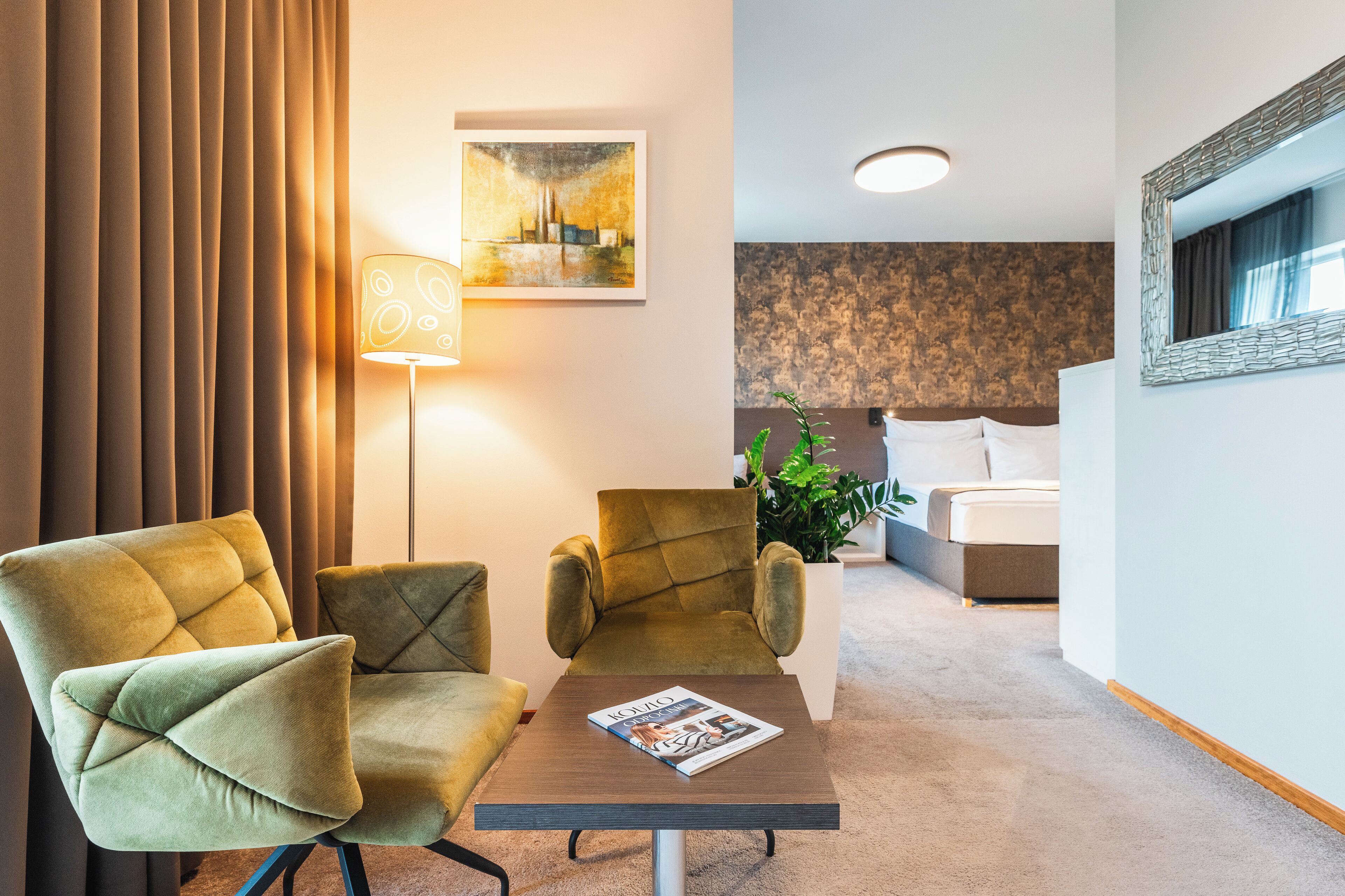 Habitación de hotel con sofá, una mesa con una revista, lámpara, un cuadro, cama y planta en maceta.