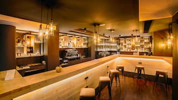 A bar area with wooden bar counters, stools, hanging light fixtures, and shelves stocked with bottles.