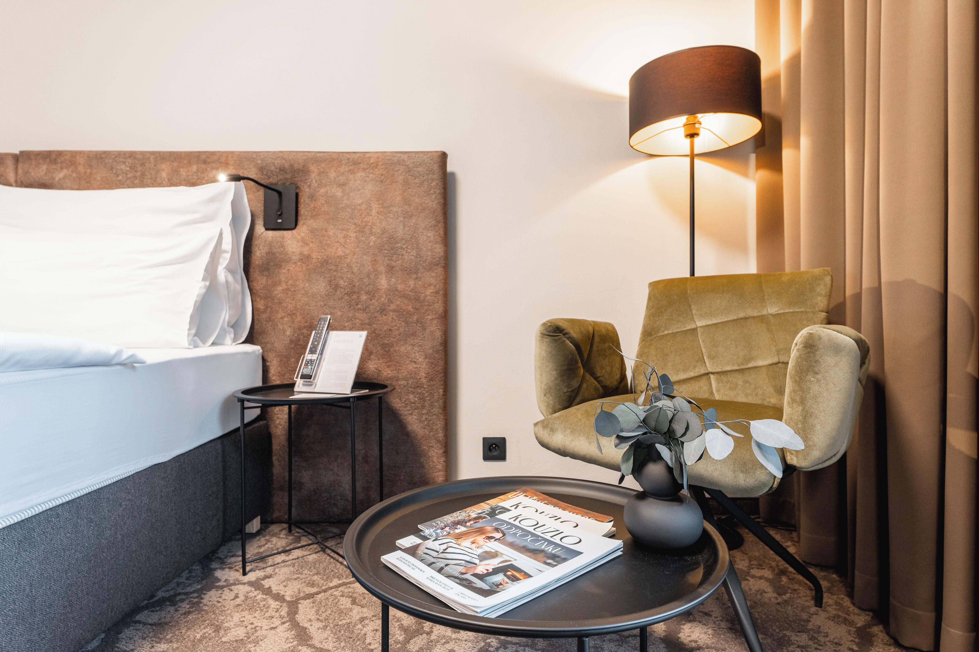 A hotel room with a bed, a small table with a magazine, a chair, a lamp, and a wall-mounted phone.
