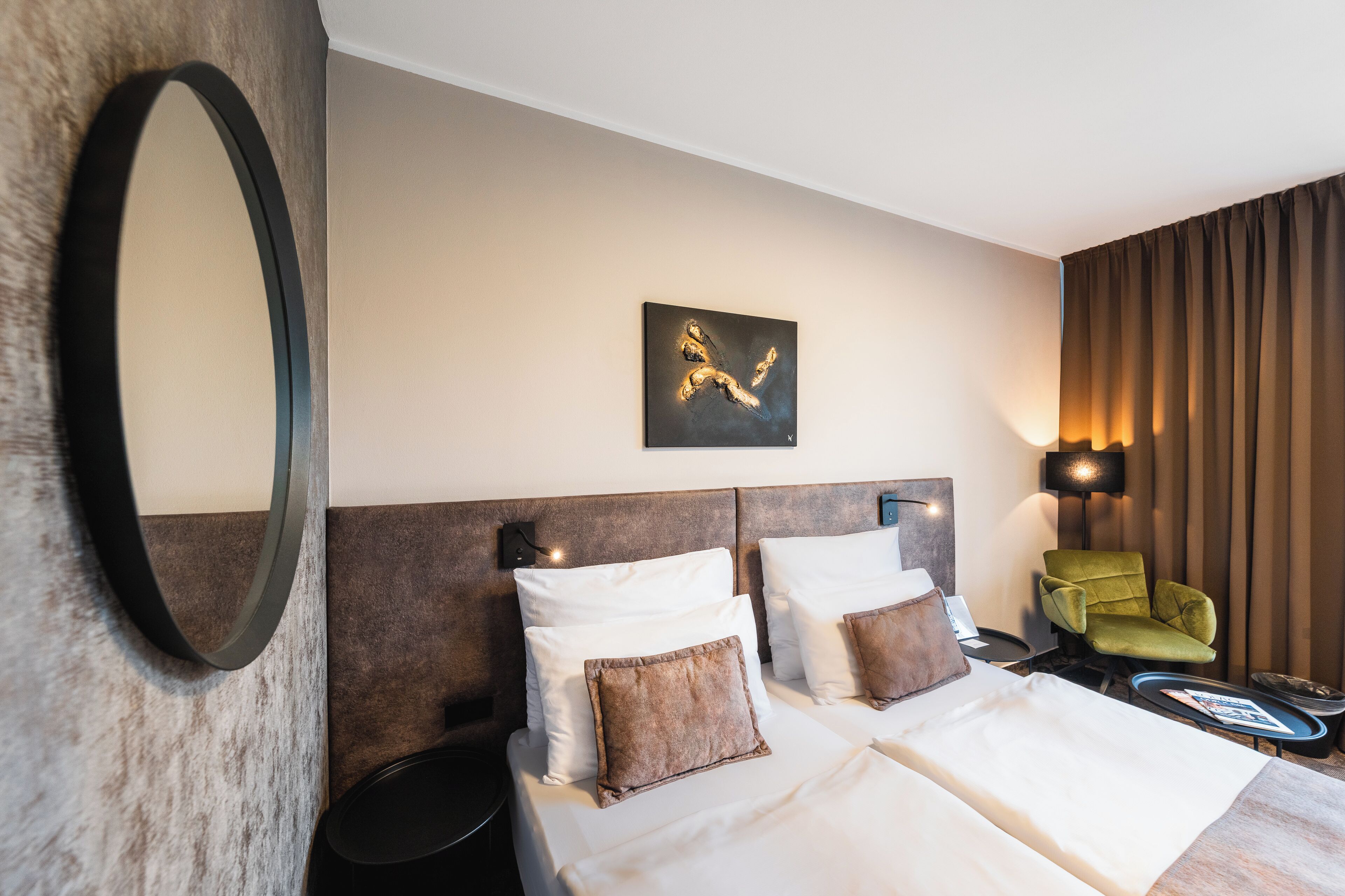A hotel room with a large bed, a headboard, a green armchair, a side table, and a wall-mounted artwork.
