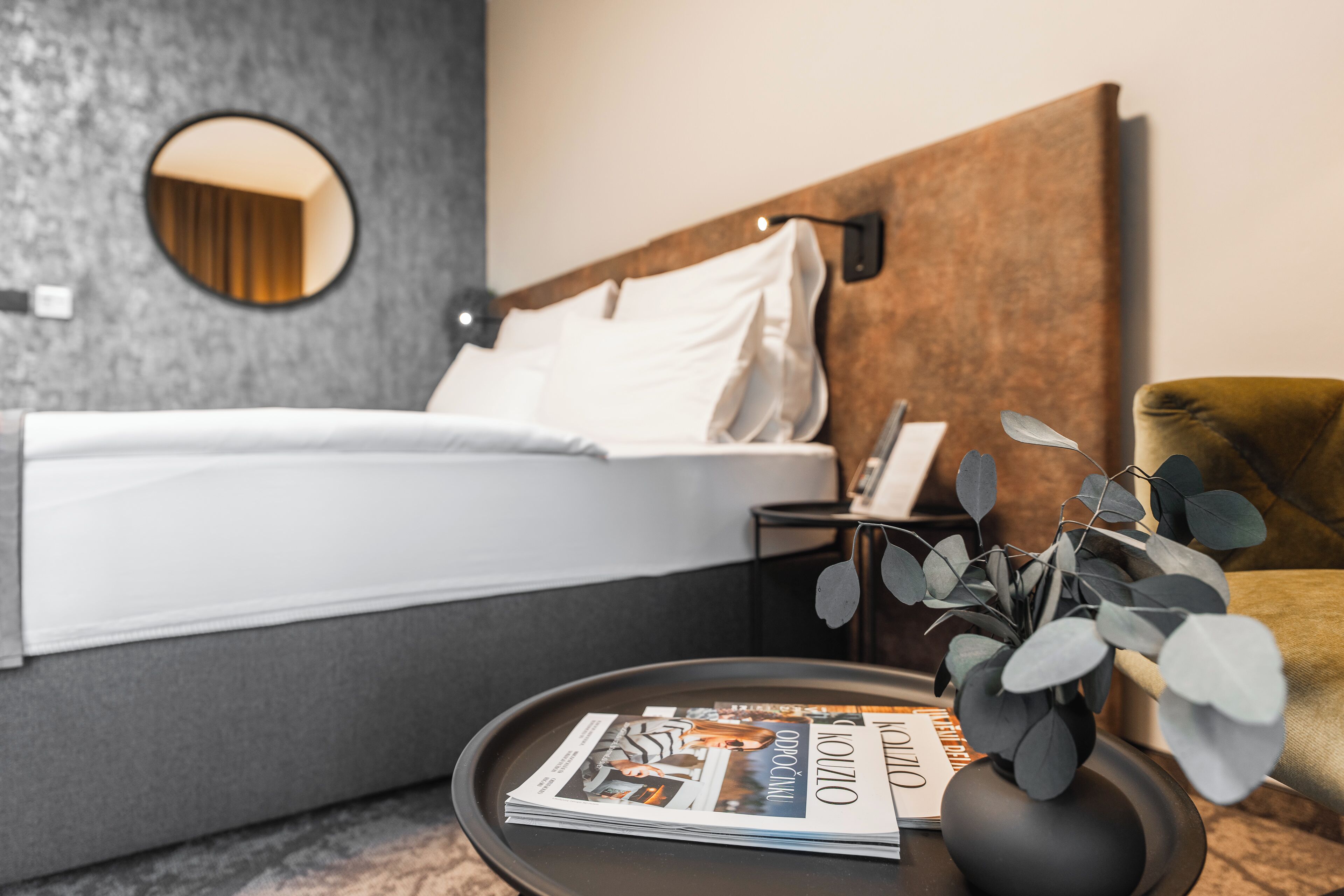 A neatly made bed with white linens, a round mirror on the wall, a bedside table with a magazine, and a green armchair.