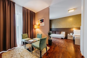 A hotel room with a wooden floor, a sofa, a dining table with chairs, a lamp, and a painting on the wall. - Maximus Resort (Brno)
