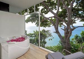 Une salle de spa avec une baignoire remplie de pétales de roses, une vue sur la mer et un grand arbre à l’extérieur.