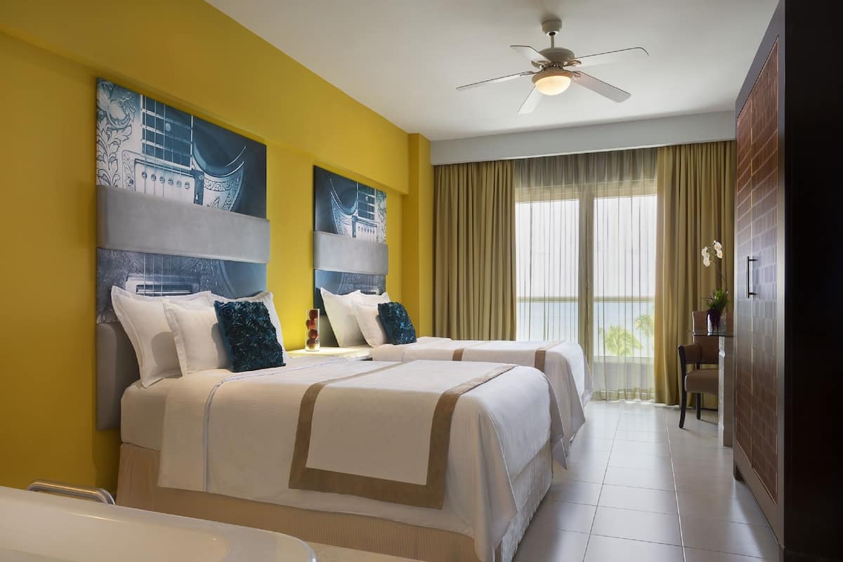 Una habitación de hotel con dos camas, un ventilador de techo, ventanas amplias con cortinas y obras de arte en las paredes.