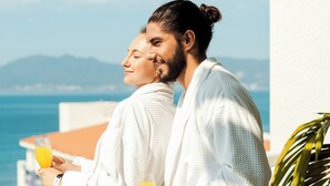 Twee mensen in witte gewaden houden glazen sinaasappelsap vast, staand op een balkon met uitzicht op zee en bergen.