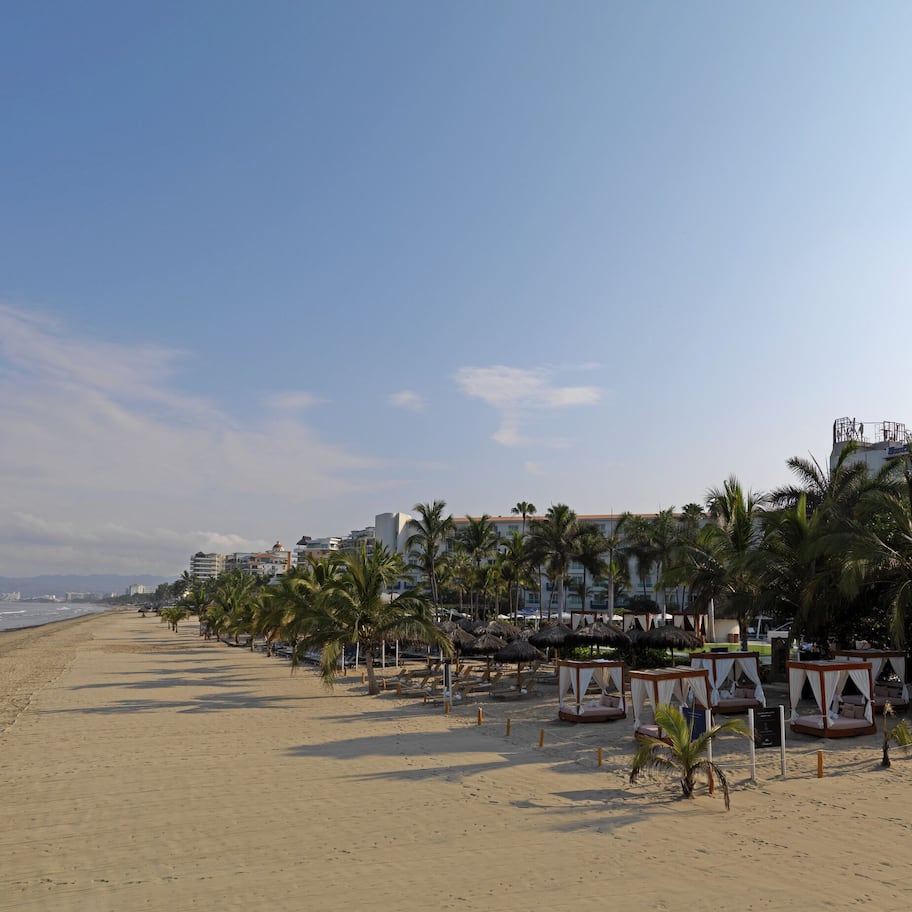 A beach with palm trees, beach huts, and a hotel in the background.
