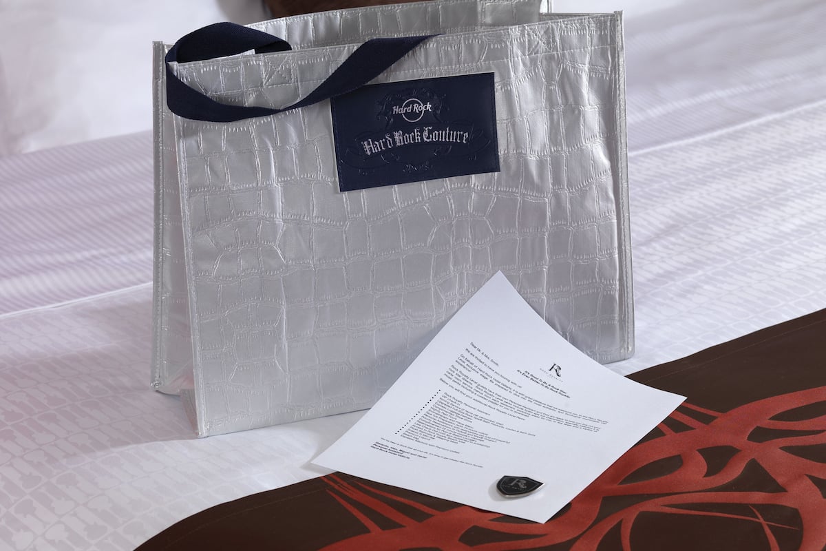 Una bolsa de regalo plateada con etiqueta negra sobre una cama con ropa de cama blanca y un cojín marrón.