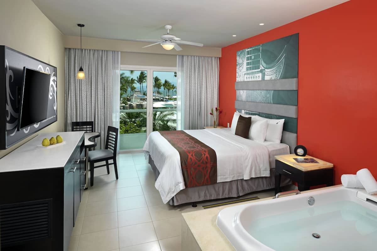 Habitación de hotel con cama, escritorio, silla, televisor, balcón con vistas y bañera.
