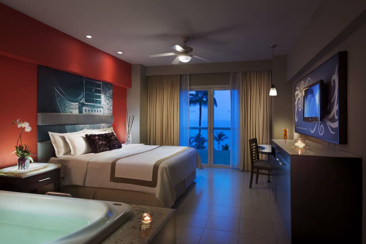 Habitación de hotel con cama, escritorio, televisor, balcón con vistas y ventilador de techo.