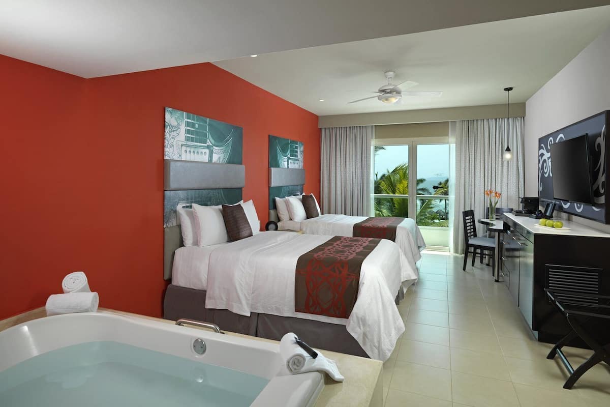 Habitación de hotel con dos camas, una bañera grande, un escritorio con televisor y un balcón con vistas.