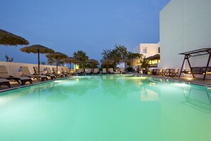 Ein Schwimmbecken mit Liegestühlen und Sonnenschirmen, umgeben von Palmen und einem Gebäude mit weißer Fassade.