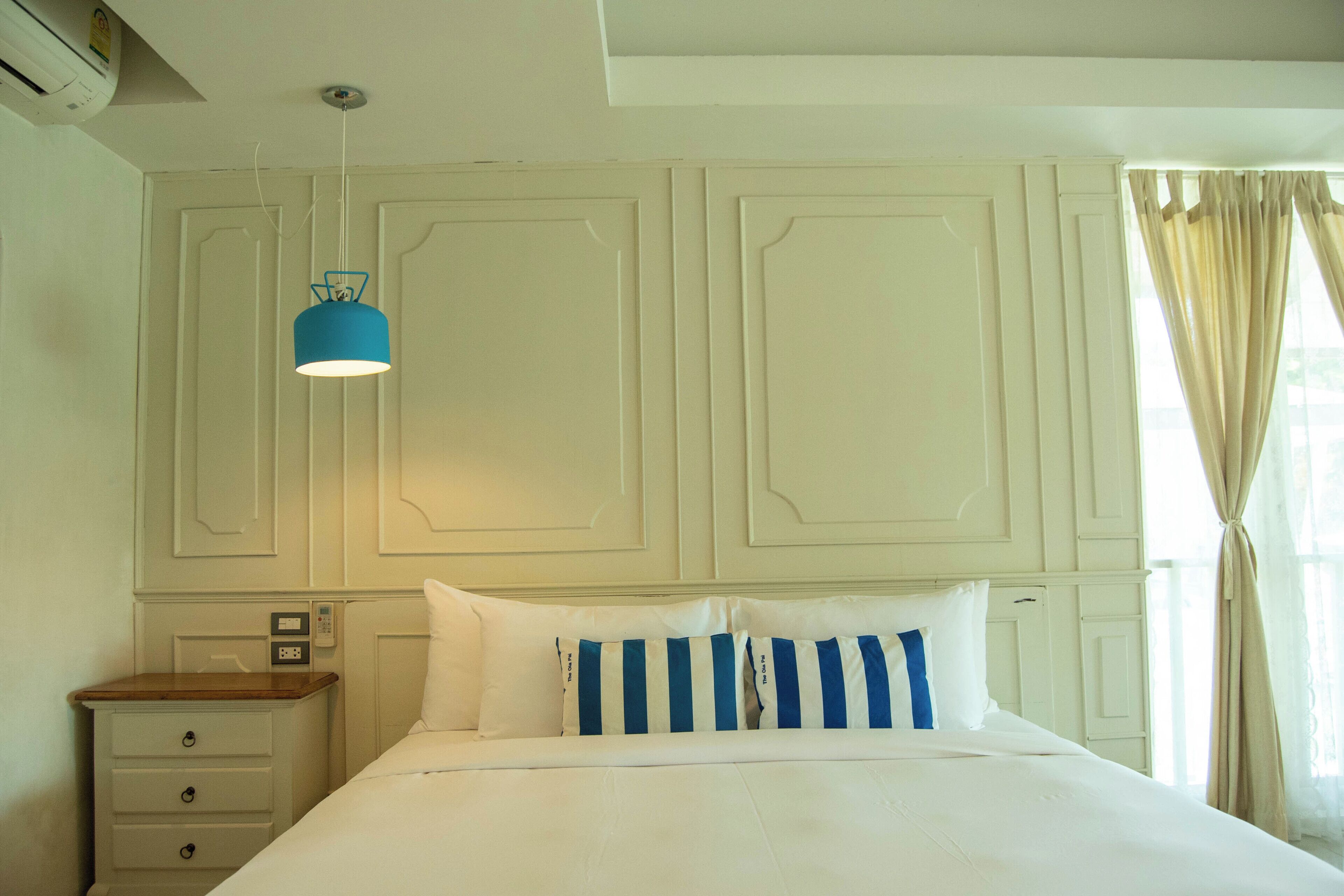 Una habitación de hotel con cama, mesita de noche, una lámpara colgante azul y una ventana con cortinas.