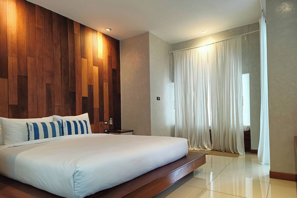 Ein modernes Hotelzimmer mit einem großen Bett, einer Wand aus Holzpaneelen und Blick ins Freie durch transparente Vorhänge.