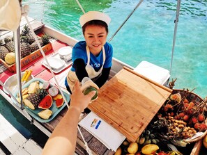 A boat with a person wearing a hat and apron, surrounded by various fruits and vegetables.