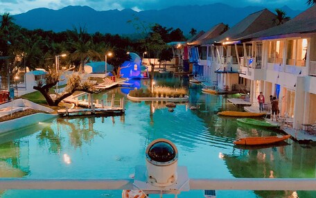 A resort pool area with a sign reading "The Oia Pai," surrounded by overwater bungalows and palm trees.