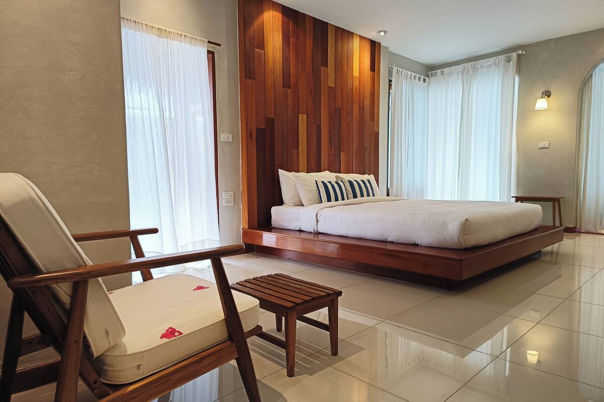 Ein modernes Hotelzimmer mit einem großen Bett, einem Holzkopfteil, einer Sitzecke mit einem Sessel und einem kleinen Tisch sowie Blick durch große Fenster nach draußen.