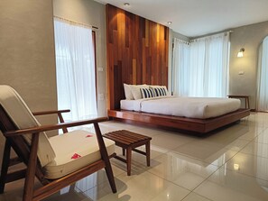 A modern hotel room with a large bed, a wooden headboard, a sitting area with a chair and a small table, and a view of the outside through large windows.