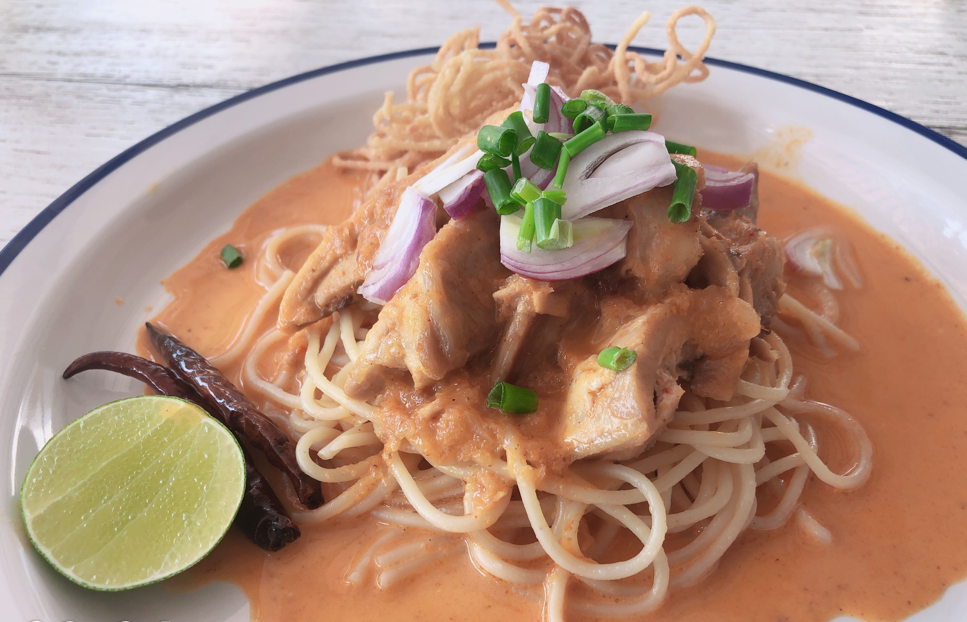 Un plato de fideos con pollo, cebolla roja, cebolletas y un gajo de lima.