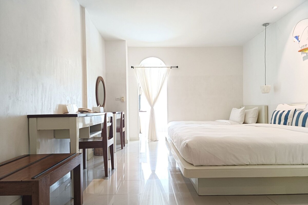 A bedroom with a bed, bedside table, chair, desk, mirror, and a window with curtains.