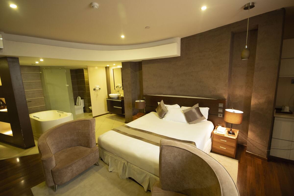 A modern hotel room with a large bed, two armchairs, a vanity with a mirror, and a bathtub.
