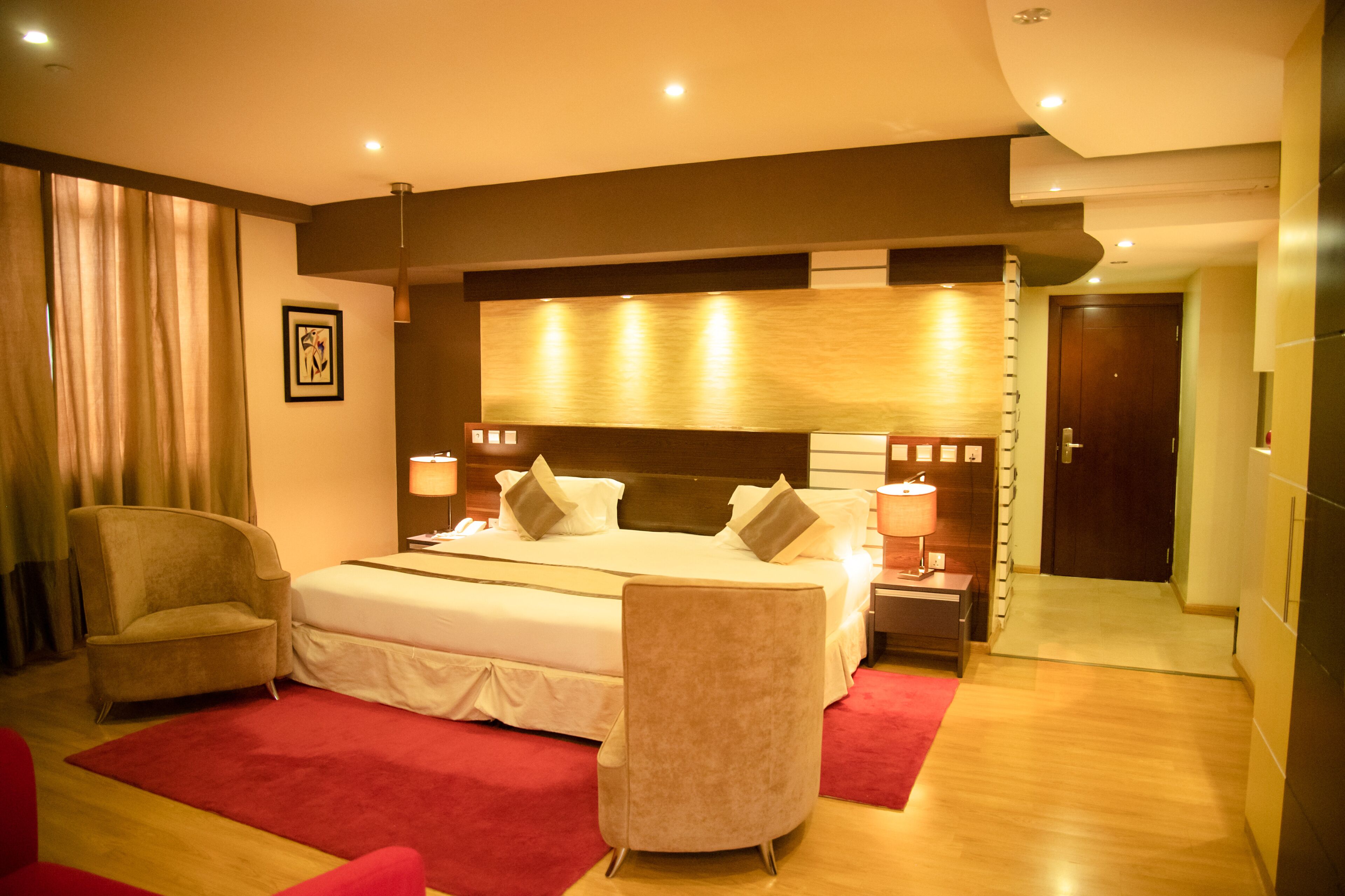 A hotel room with a large bed, two armchairs, a bedside table with lamps, and a red carpet.
