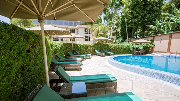A poolside area with lounge chairs and umbrellas, surrounded by greenery and a building in the background. - SG Premium Resort (Arusha)