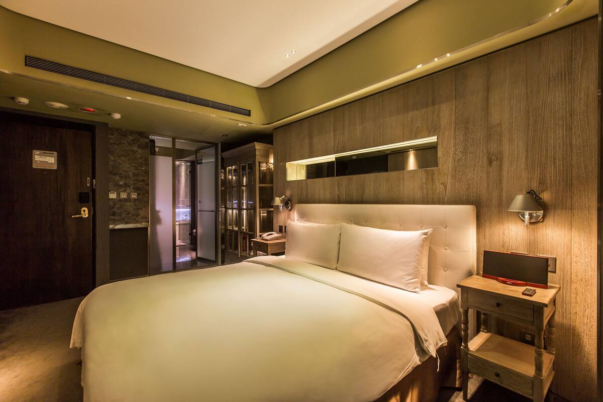 A modern hotel room with a large bed, a bedside table with a lamp, a built-in TV, and a wardrobe.