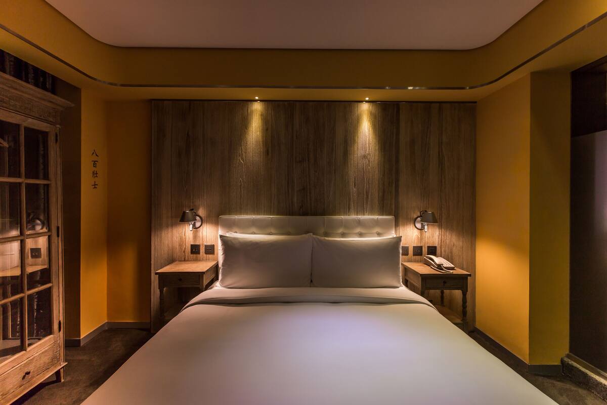 A modern hotel room with a large bed, wooden headboard, bedside lamps, and a nightstand.