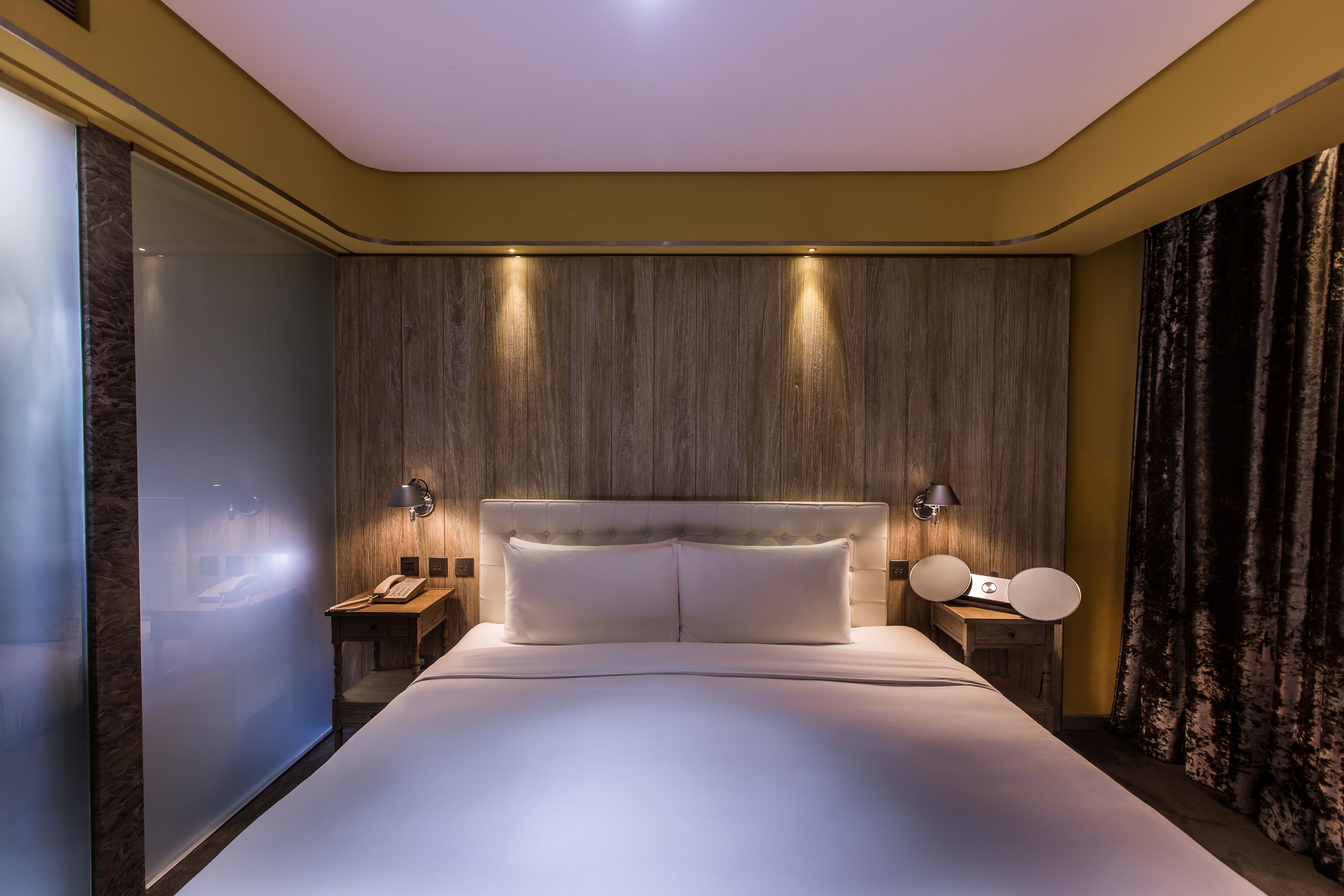 A modern hotel room with a large bed, wooden panel walls, and a nightstand with a lamp.