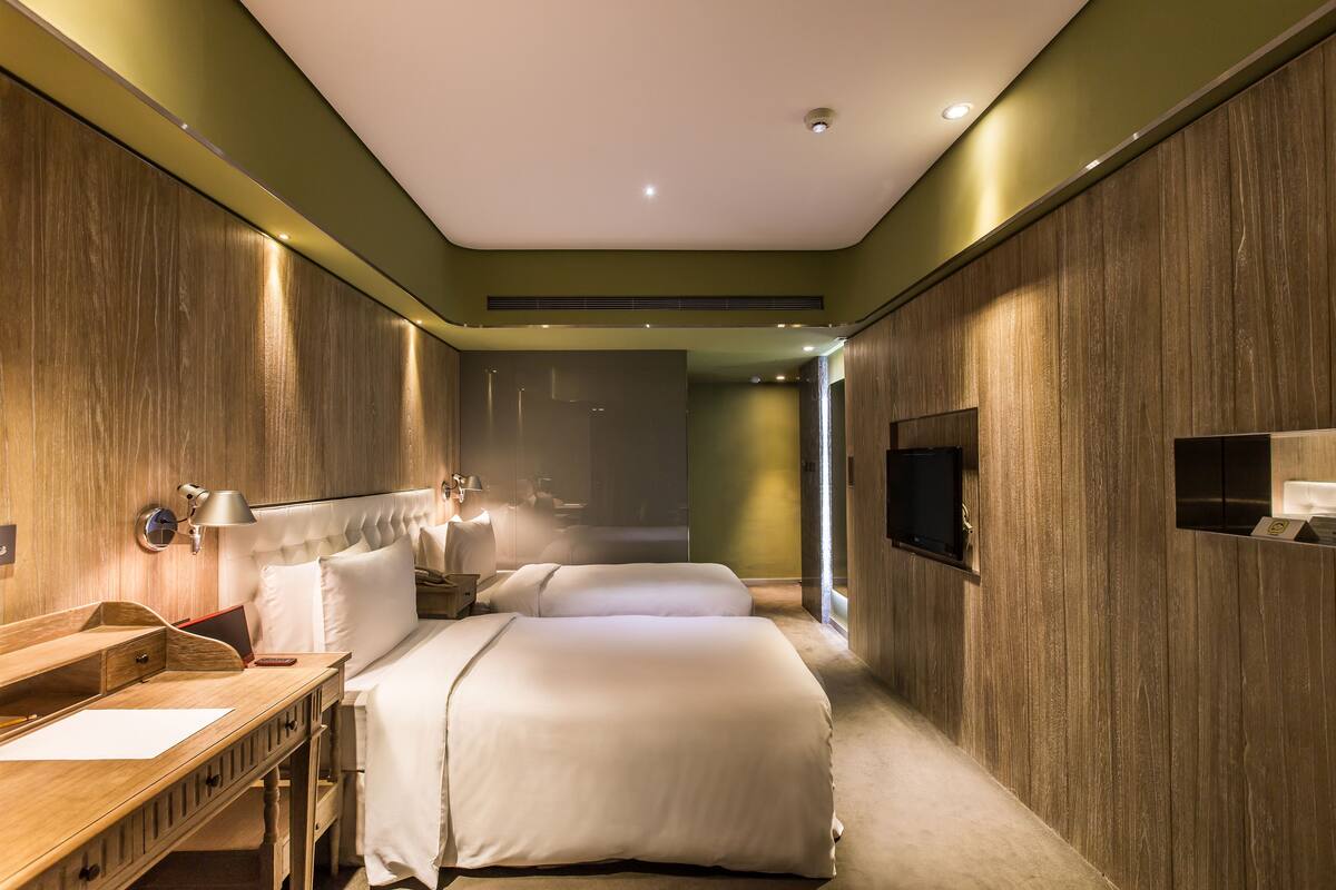 A modern hotel room with a wooden accent wall, a desk with a lamp, two beds, and a TV mounted on the wall.