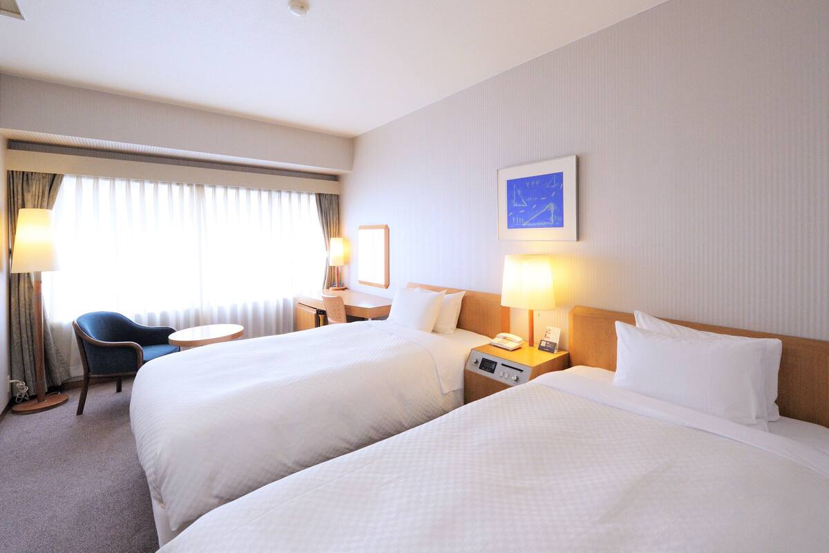Habitación de hotel con dos camas, un escritorio, una silla, una lámpara y una ventana con cortinas.