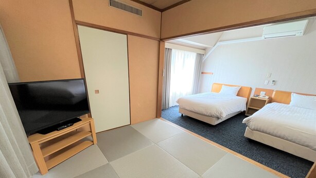 Habitación de hotel con televisión de pantalla plana sobre un soporte de madera, dos camas con ropa blanca, una mesita de noche con lámpara y un ventanal con cortinas.