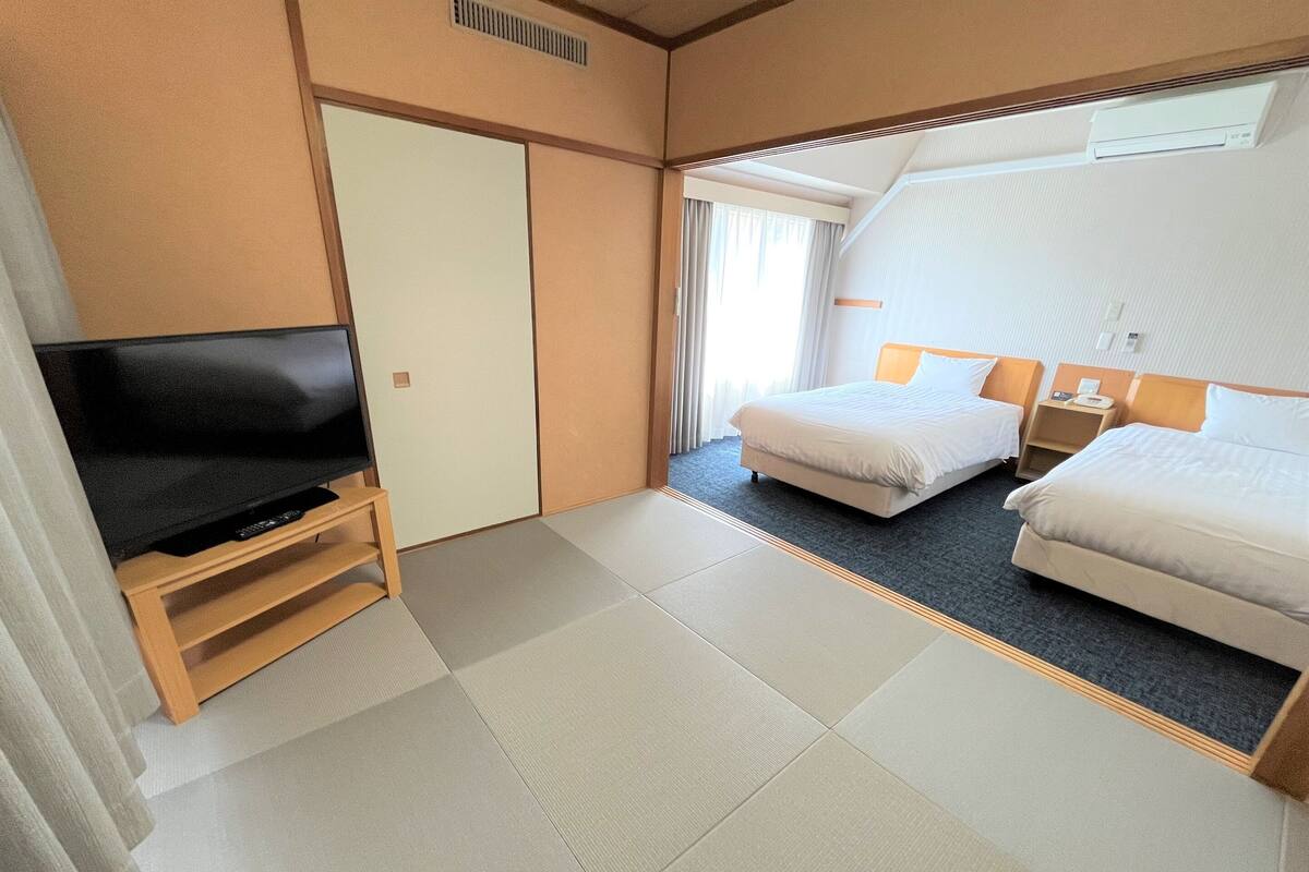 Habitación de hotel con televisión de pantalla plana sobre un soporte de madera, dos camas con ropa blanca, una mesita de noche con lámpara y un ventanal con cortinas.