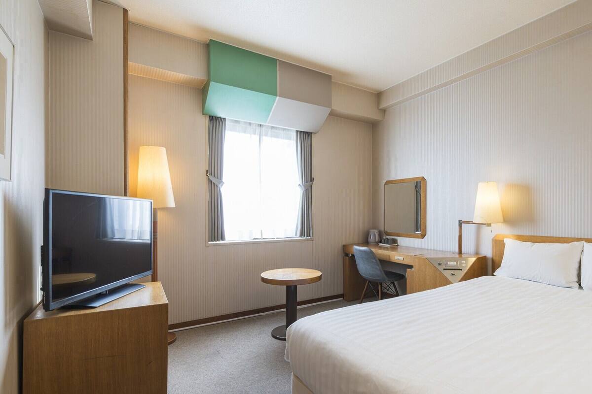 Habitación de hotel con cama, escritorio con silla, televisor, ventana con cortinas y espejo.