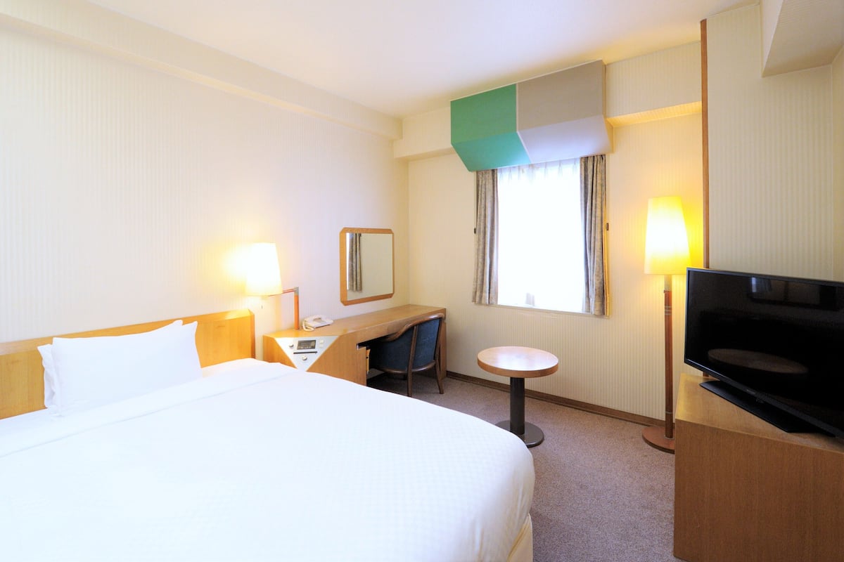Habitación de hotel con cama, escritorio, silla, televisor y ventana con cortinas.