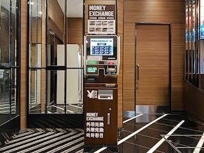 Un distributeur de devises doté d’une interface numérique, situé dans un hall moderne avec des panneaux de bois et un sol réfléchissant.
