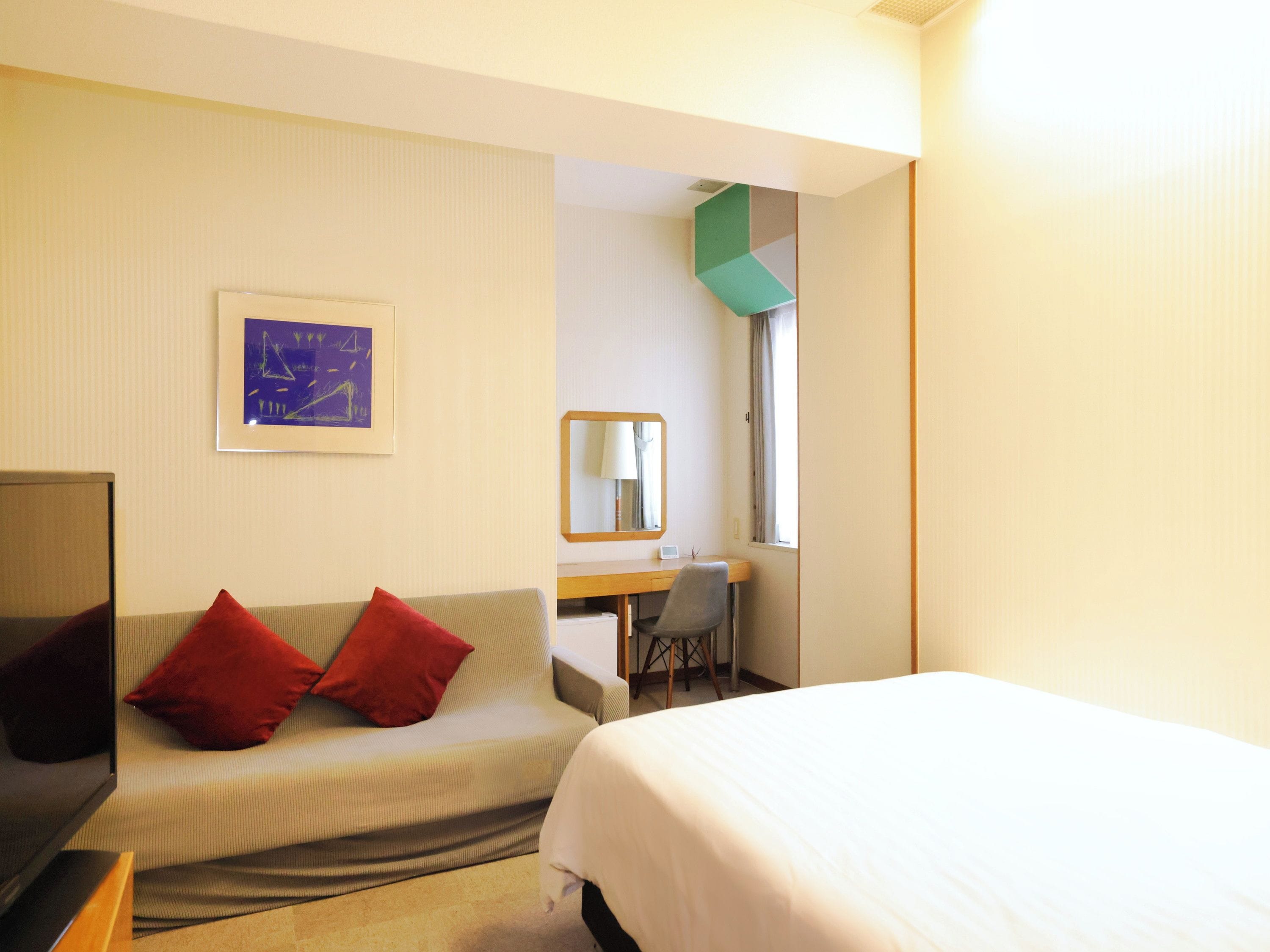 Une chambre d’hôtel avec un lit, un bureau, une chaise, une télévision et un canapé avec des coussins rouges.