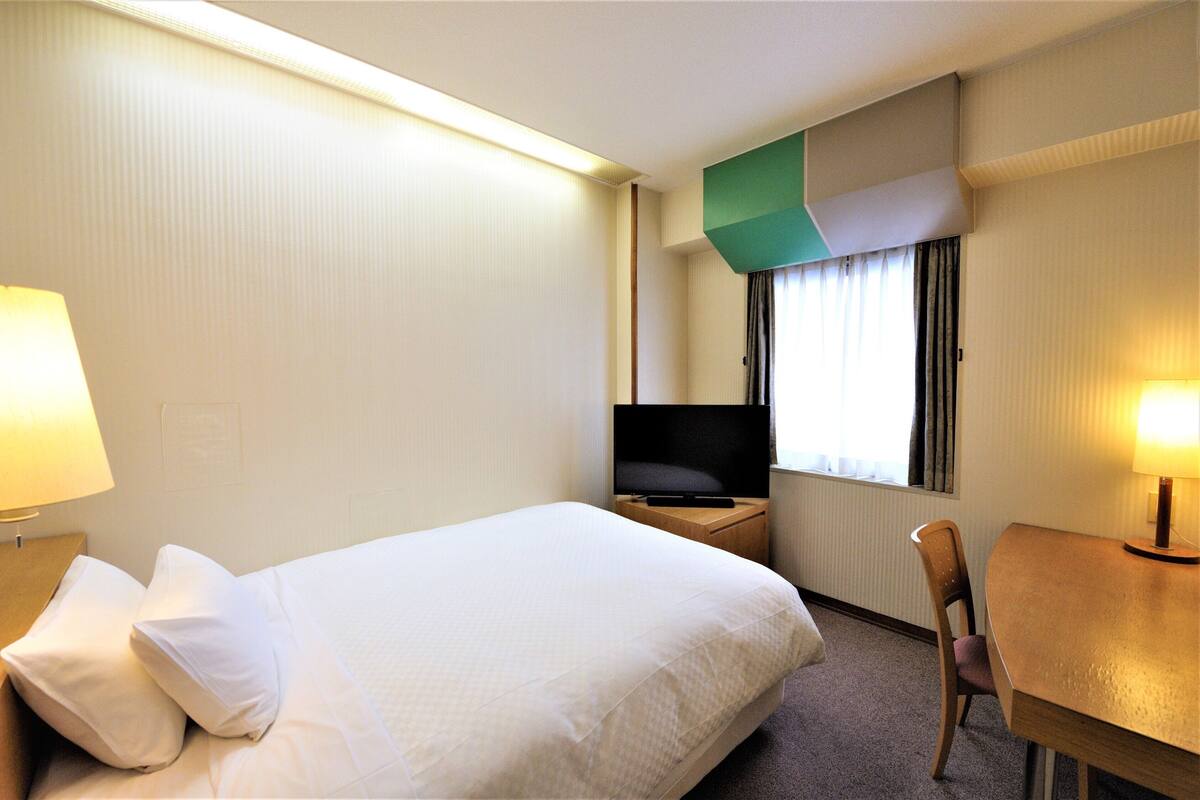 Una habitación de hotel con una cama, un televisor sobre una mesita de noche de madera, un escritorio con una silla y una ventana con cortinas.