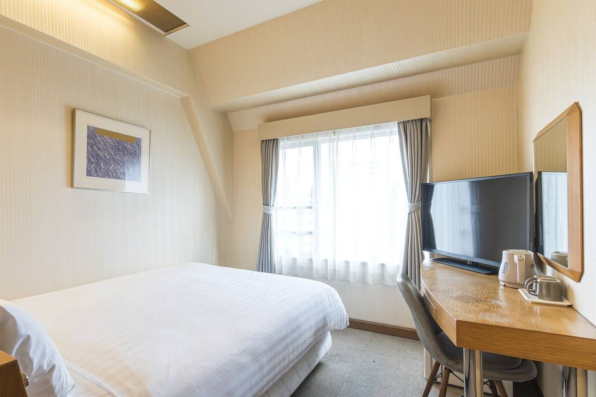 Habitación de hotel con cama, escritorio con televisor, lámpara y ventana con cortinas.